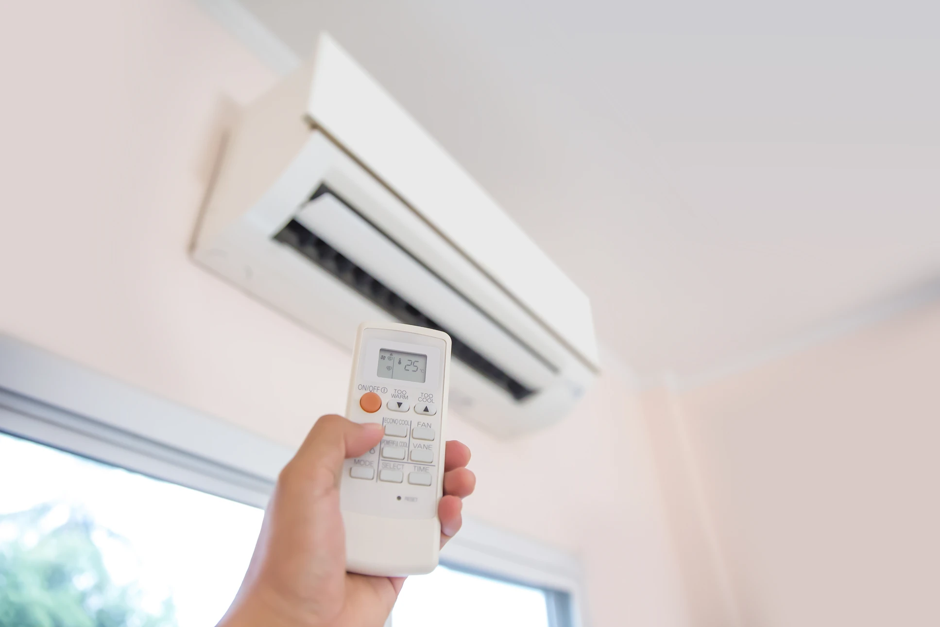 Main tenant une télécommande blanche dirigée vers une unité de climatisation murale split installée au plafond d'un intérieur résidentiel